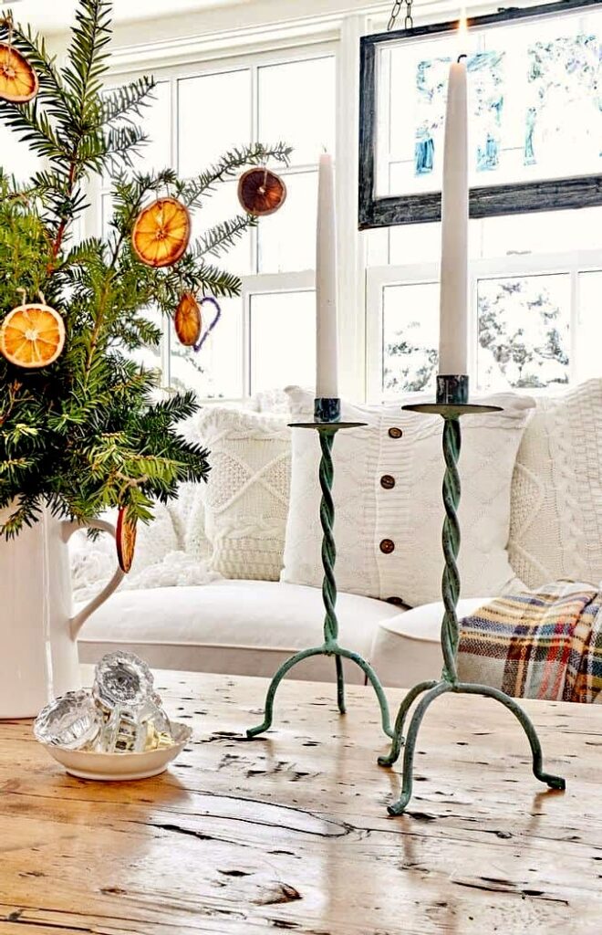 Two Patina candlesticks with white candles and Christmas oranges on coffee table with white pillows and snowy windows in the background.