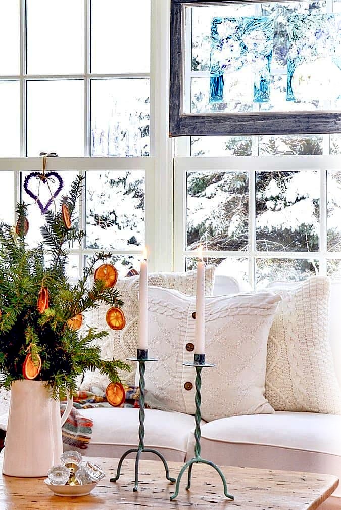 Winter decor in sunroom with cozy white pillows and pitcher of evergreens and dried oranges on coffee table.  Snowy window scene in background.