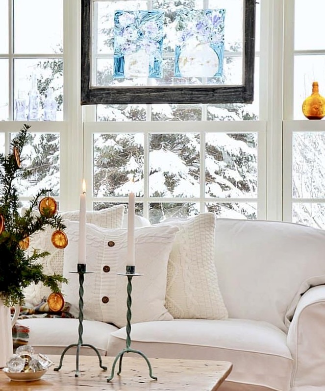 Winter white pillows on white couch next to snowy window with patina candles and evergreens and oranges on coffee table.