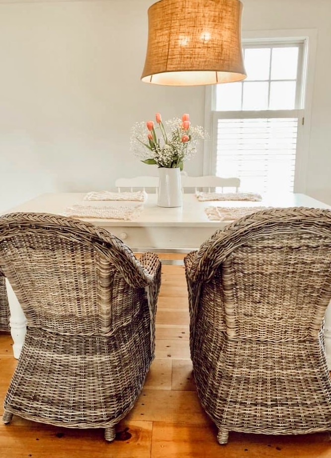 Dining room with wicker chairs white farmhouse table, pink flower arrangement in Sherwin Williams Navajo painted dining room with overhead hanging brown light.