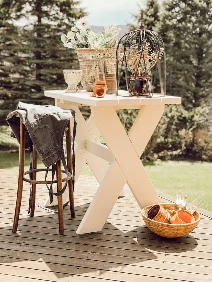 White DIY garden table with stool and apron with baskets, terracotta pots and urns outdoors on porch.