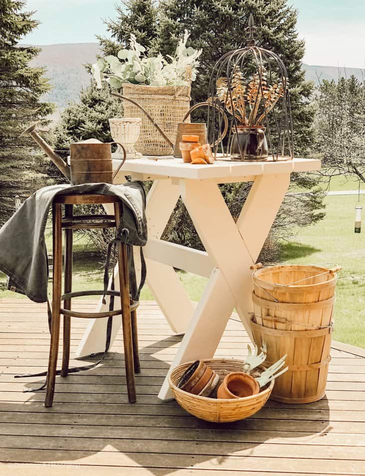 White DIY garden table with stool and apron with baskets, terracotta pots and urns outdoors on porch.