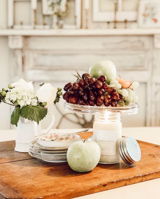 Red grapes, apples on cutting board with dishes and candle in front of vintage fireplace mantel in dining room.