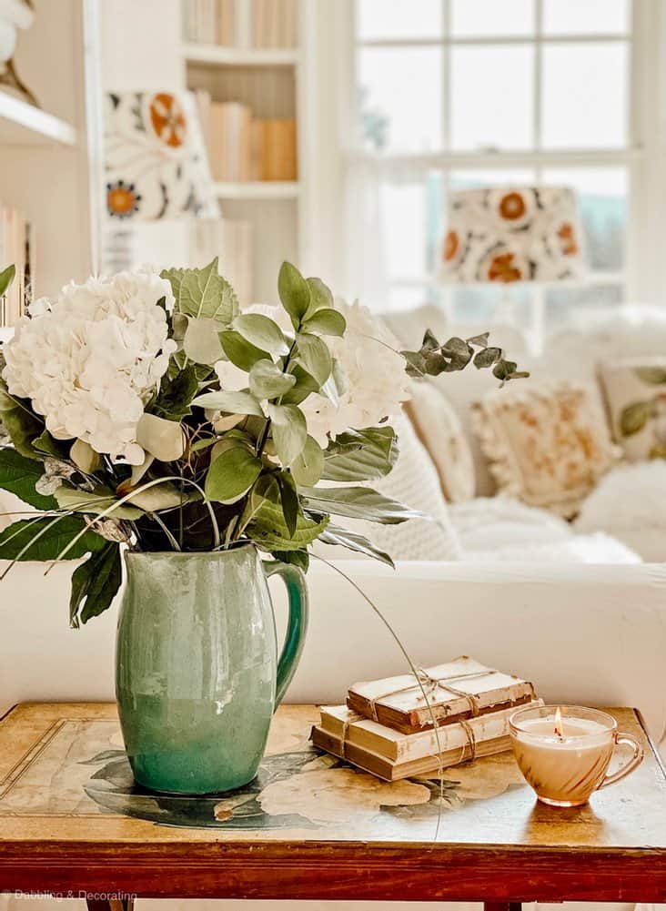 Green vase with flowers on side table with vintage winter white decorations in living room.