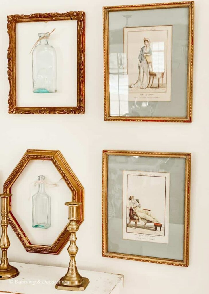 Four gold frames with bottles and pictures in them on wall over mantel with vintage brass candlesticks.