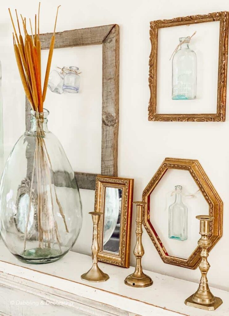 Vintage frames layered on a wall over a mantel with a large glass bottle and brass candlesticks.