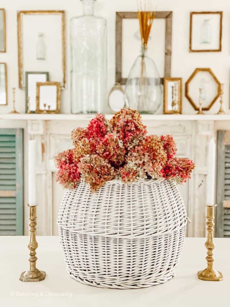 White basket of pink hydrangeas on white farmhouse table with brass candlesticks in front of vintage mantel with layered vintage frames.