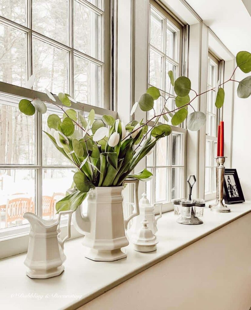 Vintage windowsill decor with white porcelain tea set with greens, candles and pictures on ledge.