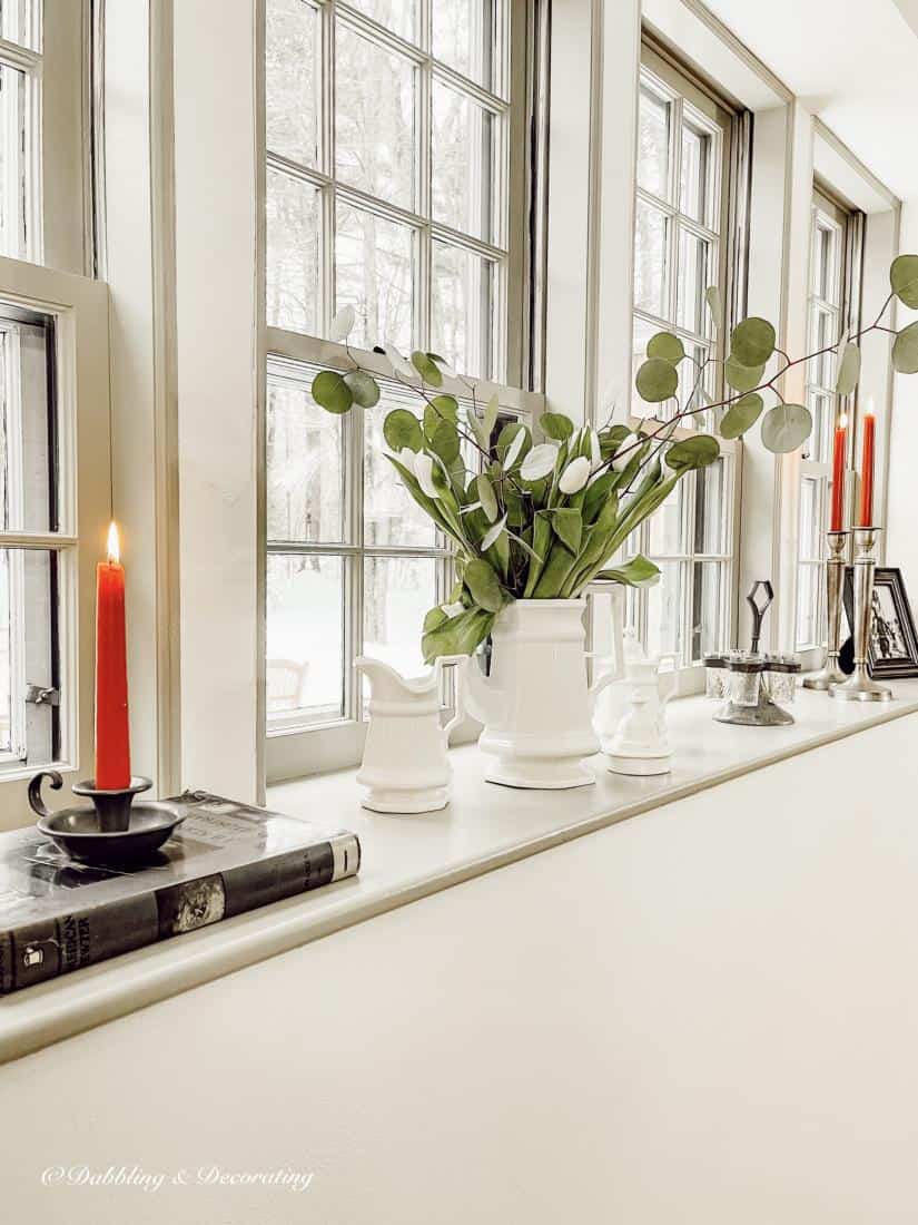 Vintage style windowsill decor with candles, books, white tea set and fresh greenery on the ledge.