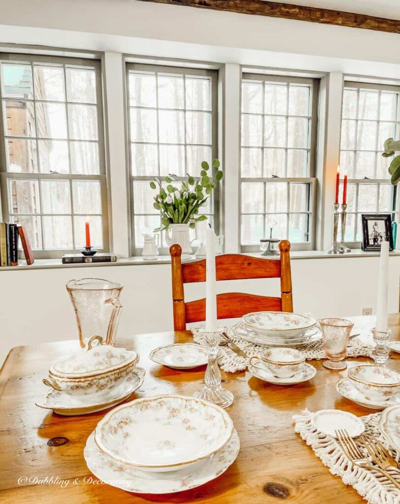 Vintage Dining room with table set with vintage dinnerware and windowsill in the background with four tall windows.