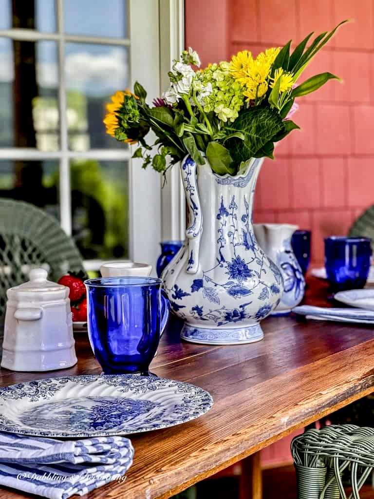 Vintage wallpaper table with blue and white vase of flowers and table setting on porch.