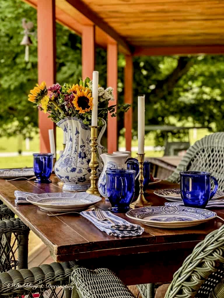 Wooden table on outdoor porch with blue and white table settings and green wicker chairs.