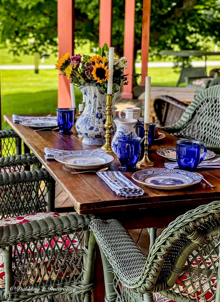 Outdoor wooden table on porch with blue and white table settings and green wicker chairs.