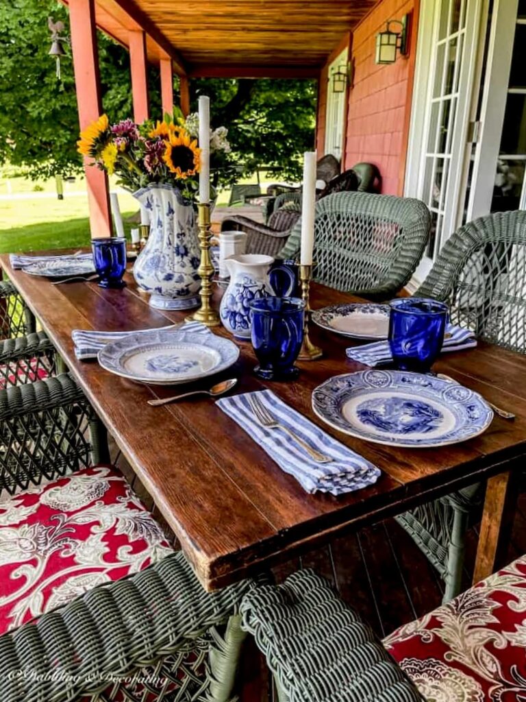 Outdoor wooden table on porch with blue and white table settings and green wicker chairs.