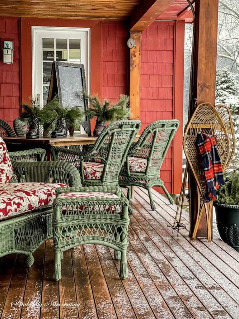 Green wicker furniture on ski house decor porch with vintage snowshoes, red scarf and evergreens in vintage ski boots.