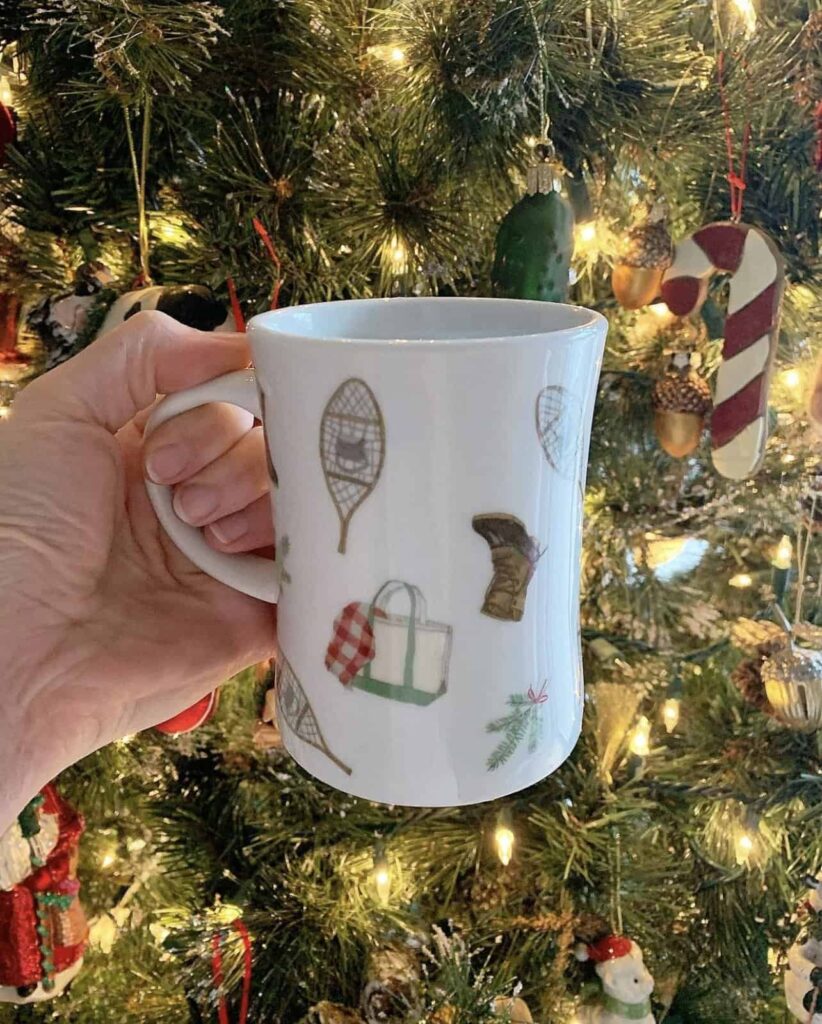 Sara Fitz mug in front of Christmas tree with LL Bean bags and boots and snowshoes design.