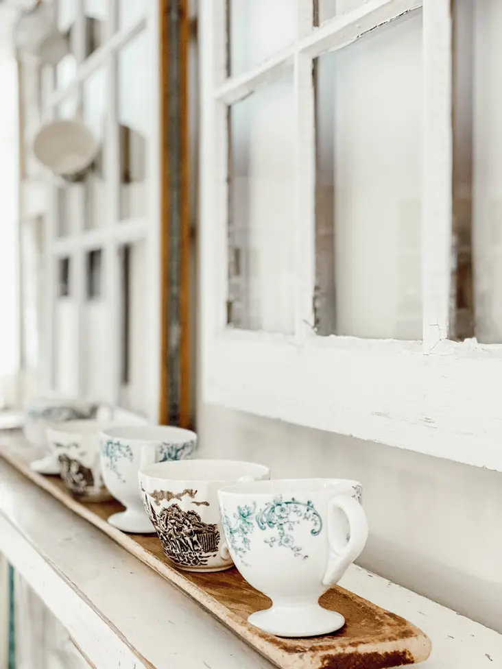 Vintage teacups on mantel with old windows.