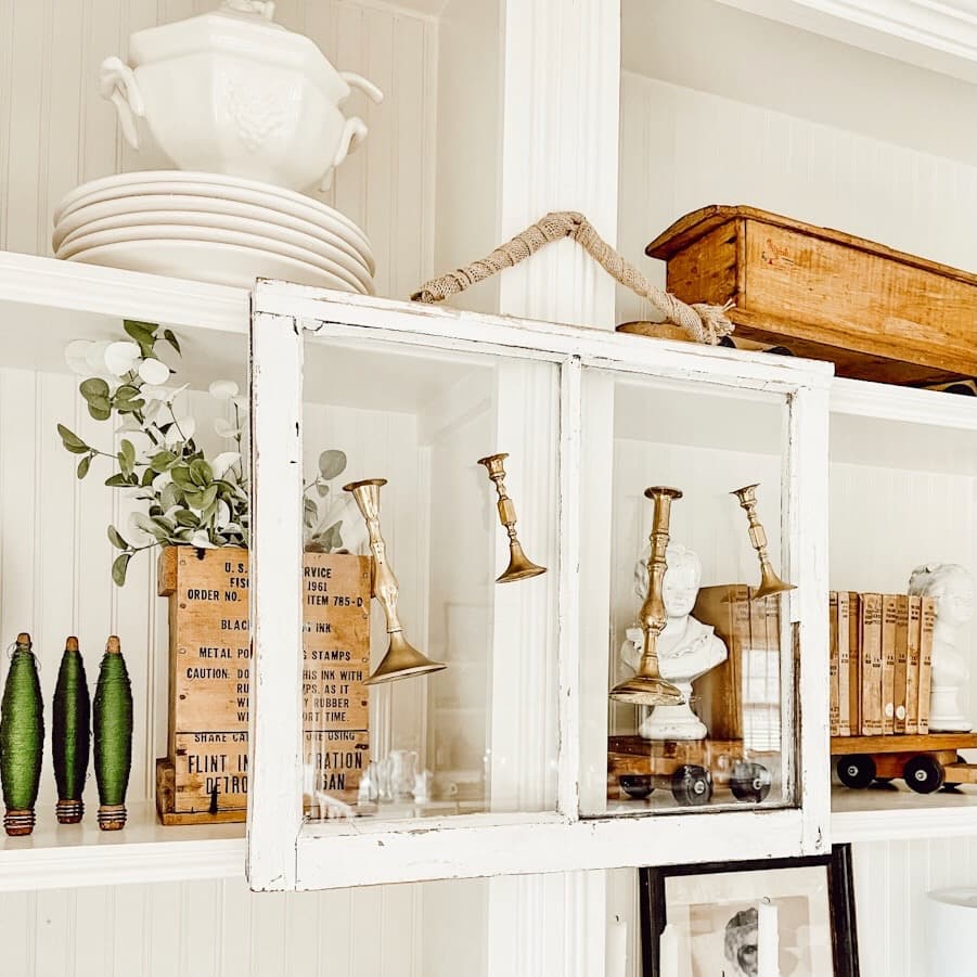 Vintage brass candlesticks hanging from a white salvaged window on vintage style bookcase.