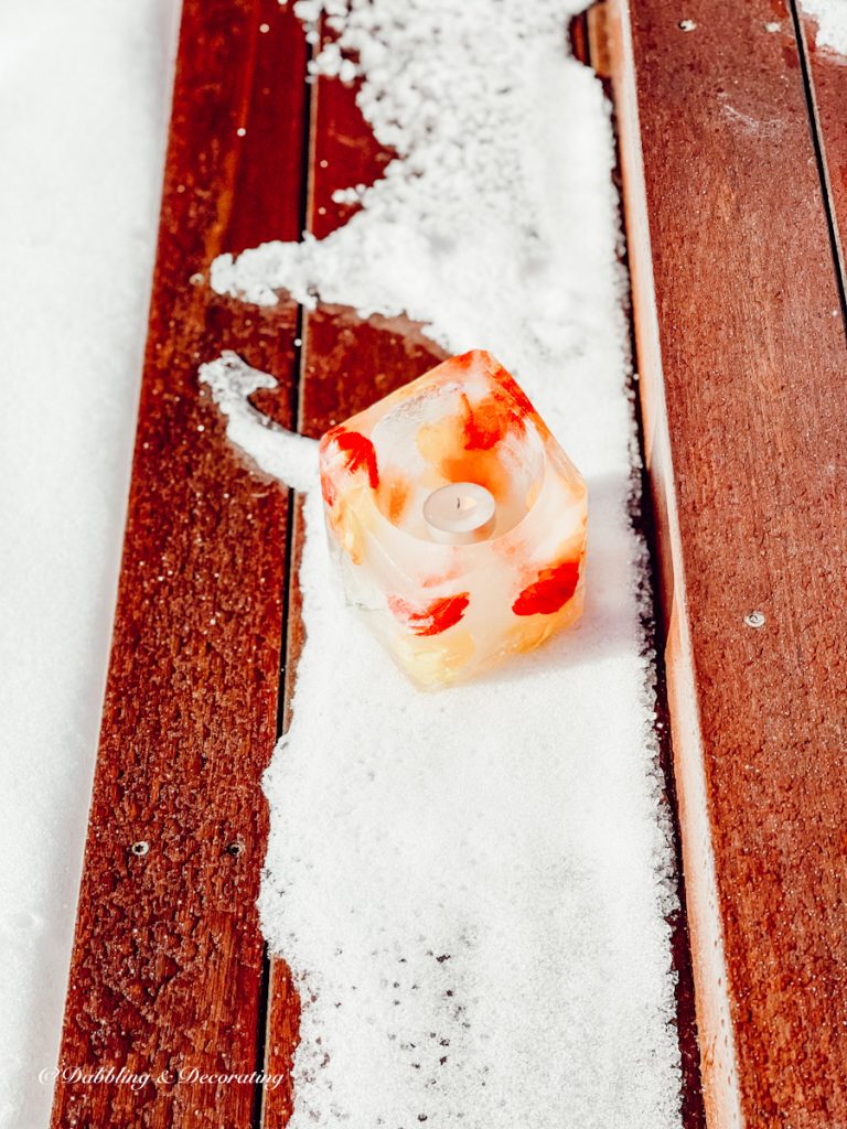 Ice Candle with red and yellow accents and tea light candle in snow on wooden porch steps.