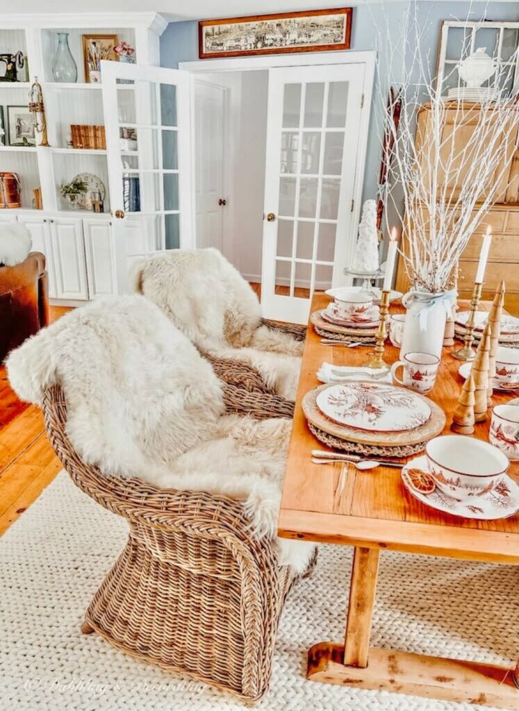 Vintage pine table with Toile Winter Wonderland dinnerware, wicker chairs with sheepskins in dining room.