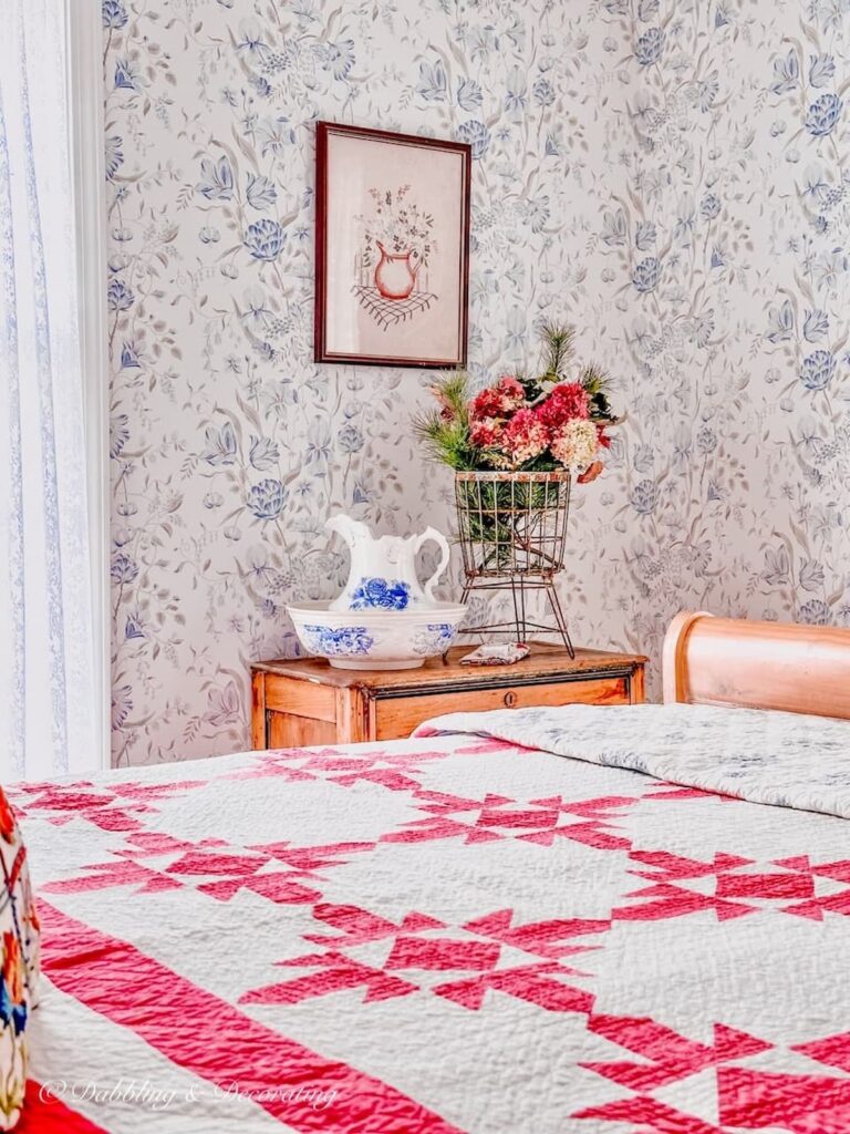 Vintage pink quilt on bed with fall flower arrangement in basket with wallpaper on wall.