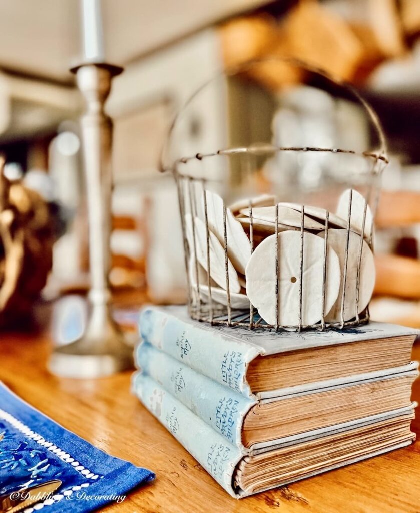 Sand dollar collection in antique wire basket on stack of blue books on table.