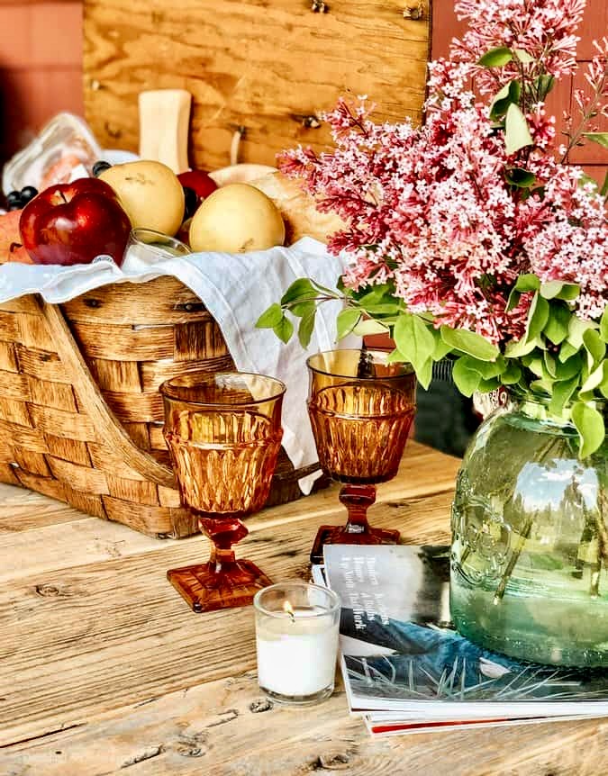 Fruit basket on table with two vintage brown glasses and jar full of lilacs.