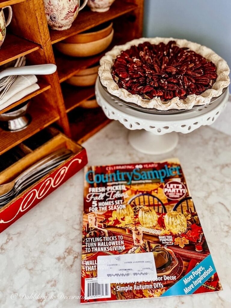 Pecan pie on vintage pie stand with Country sampler fall magazine edition on country style kitchen counter.