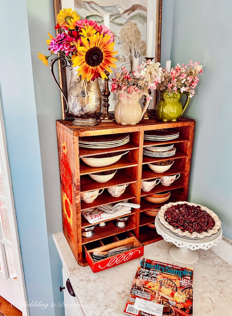 Vintage wood pie safe on kitchen counter decorated with vintage dishes and flowers.