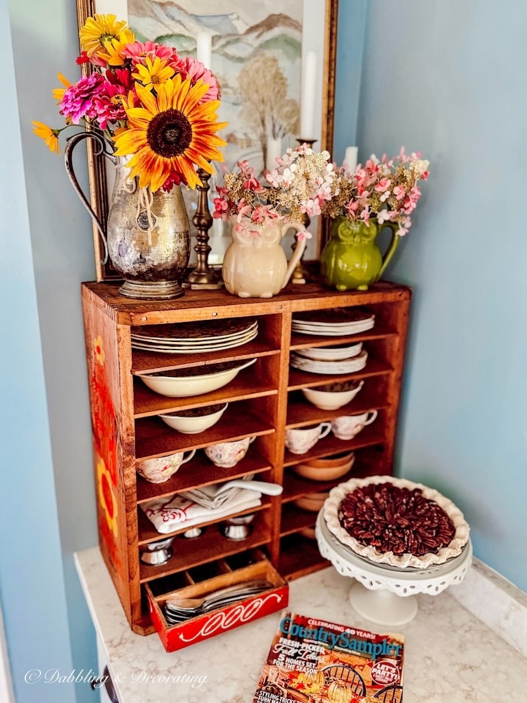 Vintage wood pie safe on kitchen counter decorated with vintage dishes and flowers.