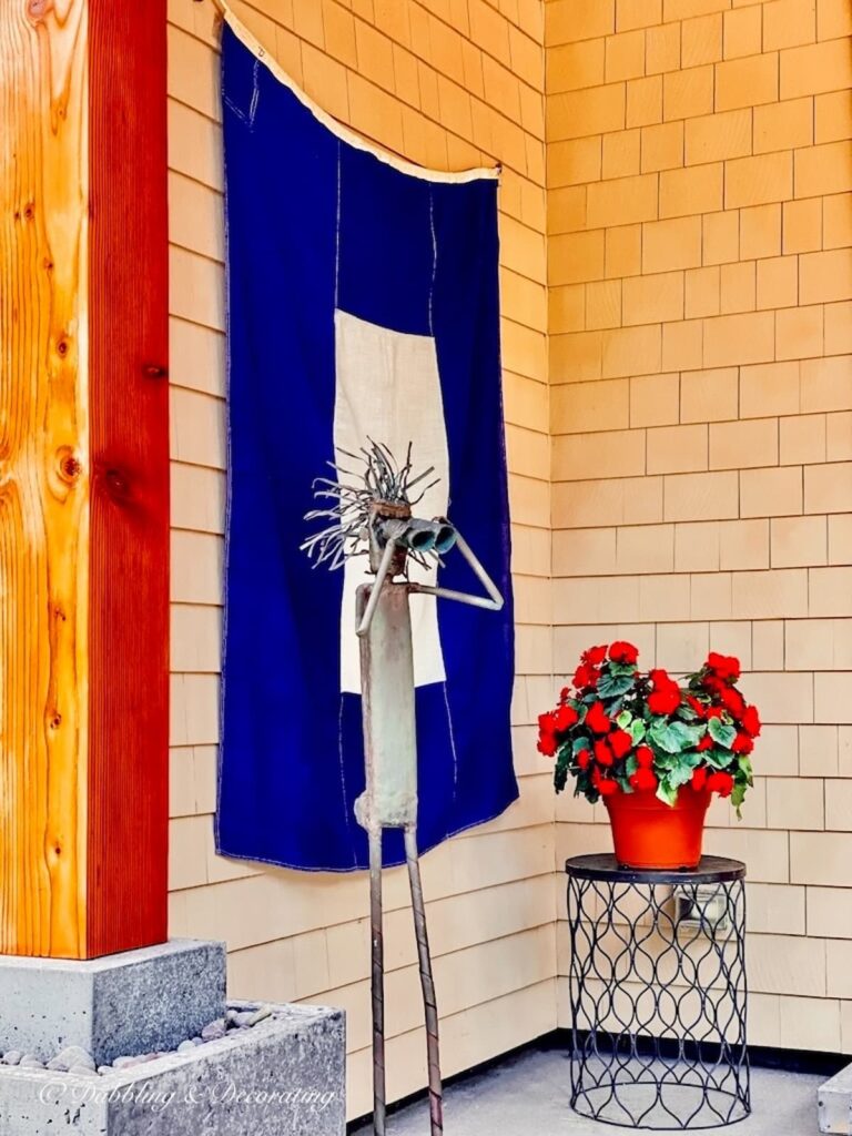 Vintage blue and white signal nautical flag hanging as welcome at Coastal Maine home with iron man with binoculars and side table with red petunias.