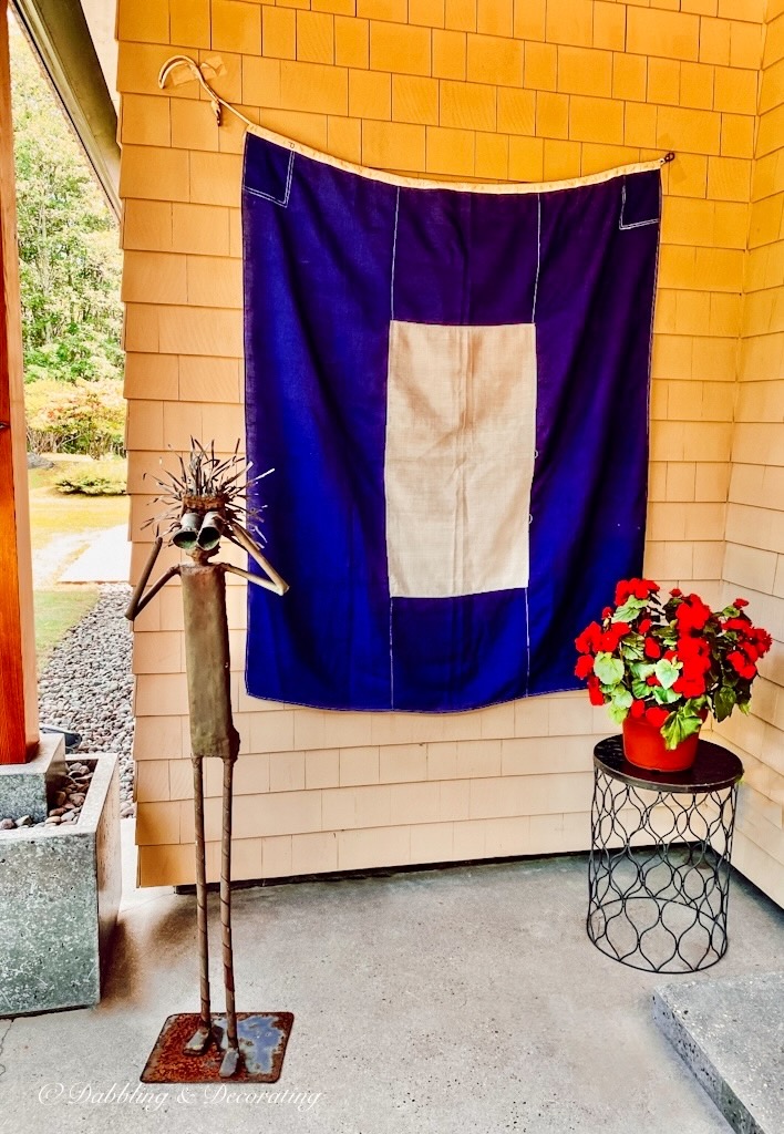 Vintage blue and white signal nautical flag hanging as welcome at Coastal Maine home with iron man with binoculars and side table with red petunias.