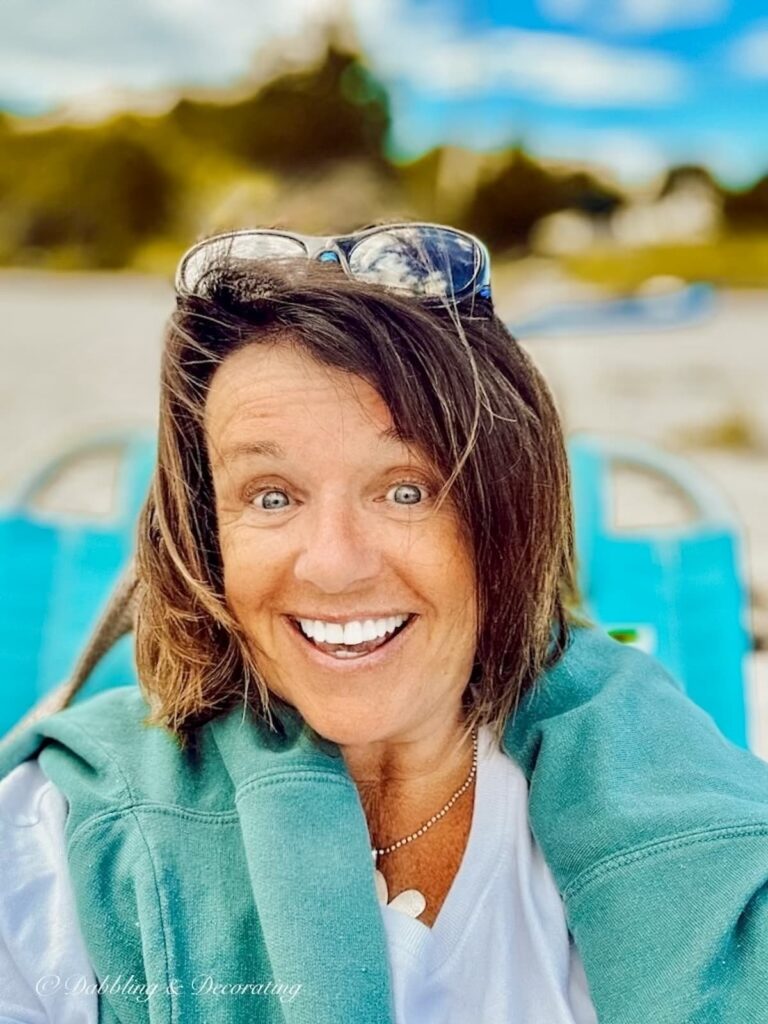 Ann, home decor blogger sitting on the beach in Maine with green sweatshirt and sunglasses on Head.