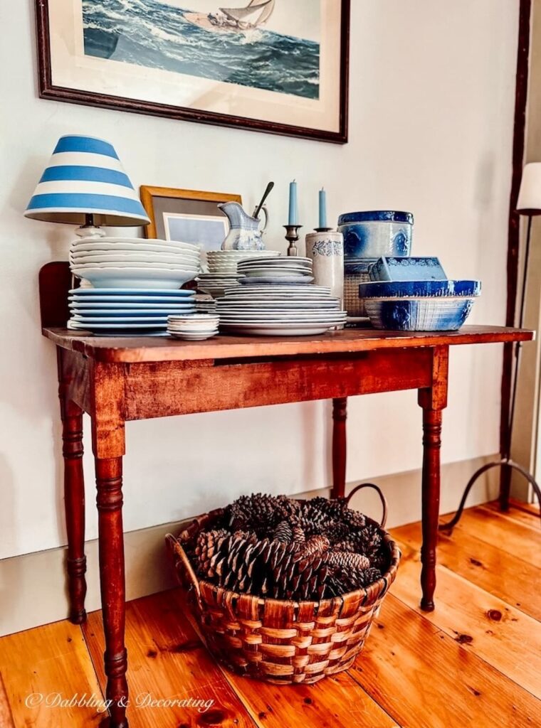 Basket of pine cones on wood flooring under antique table topped with blue and white dishes.