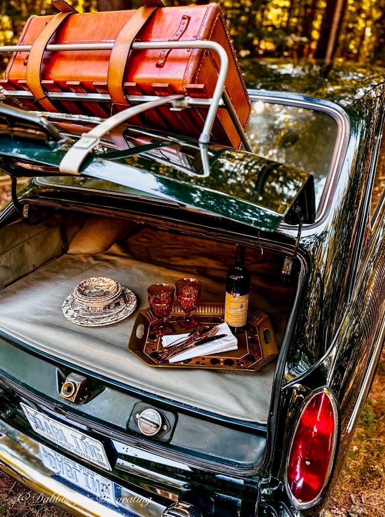 Vintage Toleware tray with bottle of wine and two Mount Vernon Amber glasses with brown and white transferware dishes in vintage car trunk.