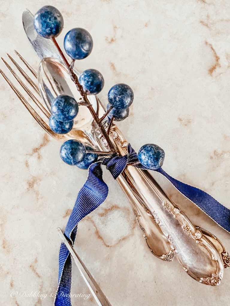 Cutting a folded blue ribbon together with a silverware place setting and a blueberry branch with a blue ribbon.