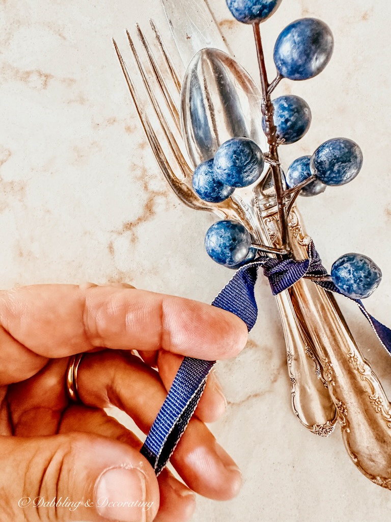 Folding a blue ribbon in hand tieing together silverware place setting and a blueberry branch with a blue ribbon.