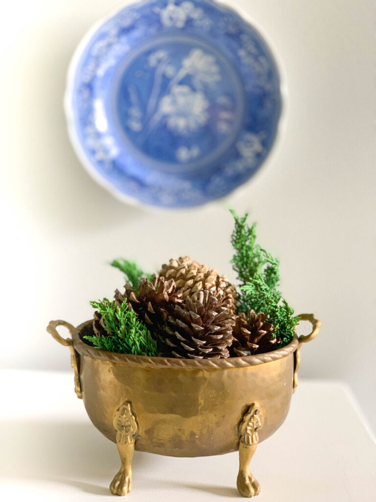 Gold vintage bowl of pinecone decor with greenery and blue plate on the wall.