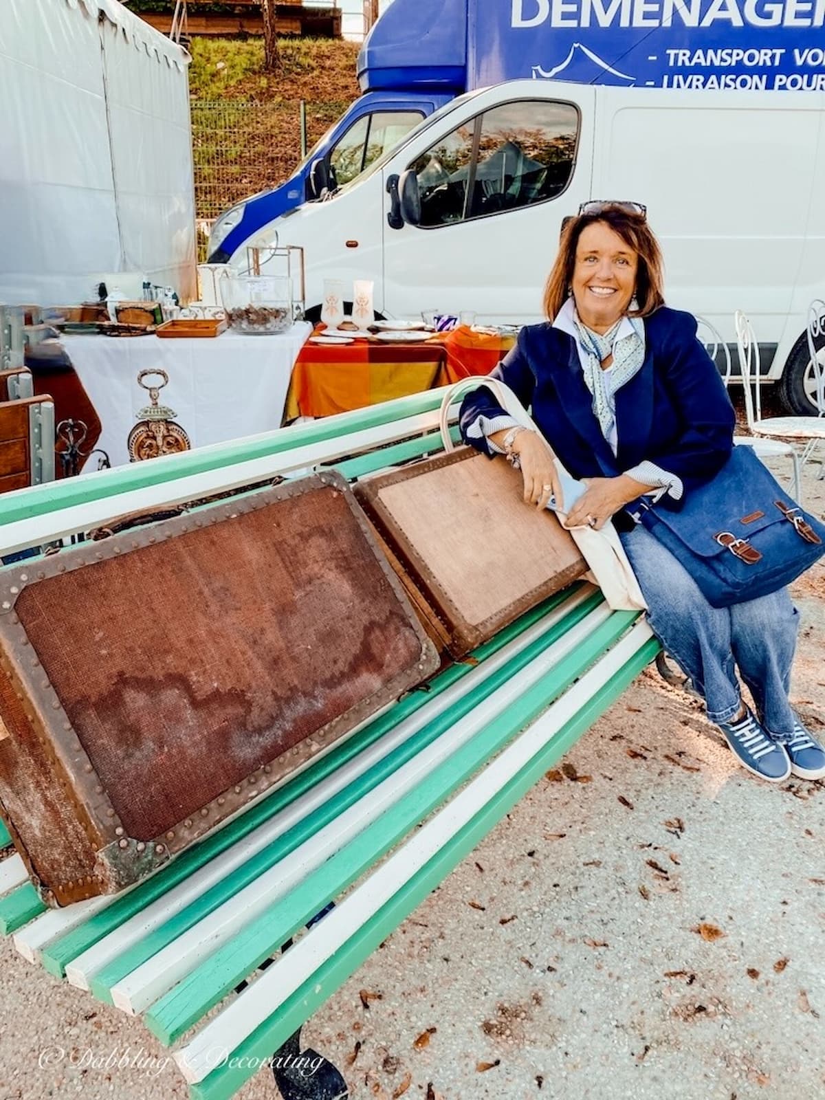 Ann, home decor blogger sitting on a green and white striped bench with vintage suitcases at a Paris flea market.