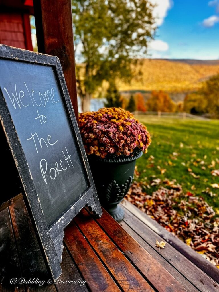 Chalkboard sign with Welcome to the Porch sign next to planter of mums with Vermont fall foliage views.