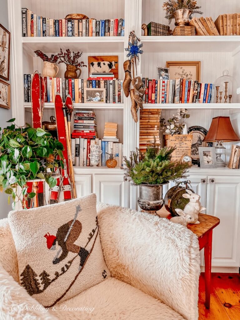 Cozy corner book nook with sheepskin chair, side table plants, and hanging decorative wood spindles on bookshelving.