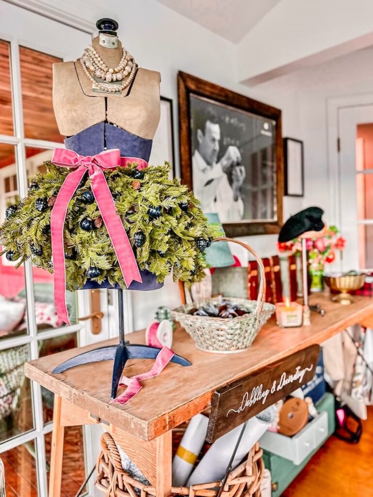 Vintage Christmas mannequin styled for the holidays with evergreens and a pink bow on a craft table in craft room.