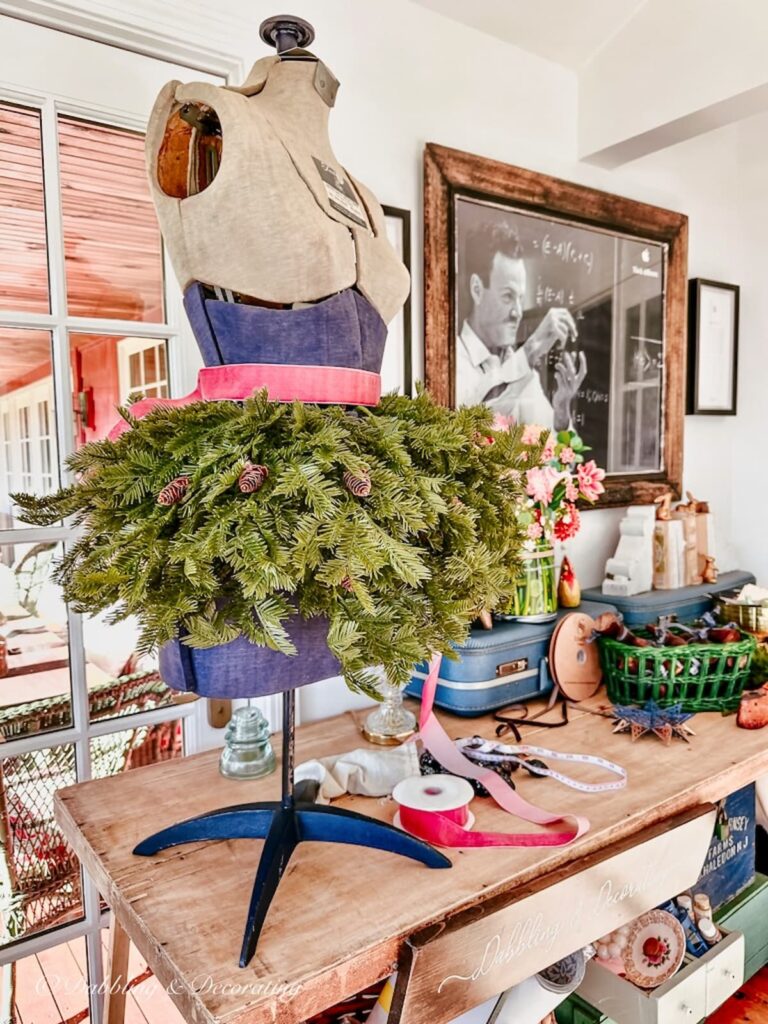 Vintage table top mannequin with evergreens around waist with pink ribbon for Christmas decor on craft table.