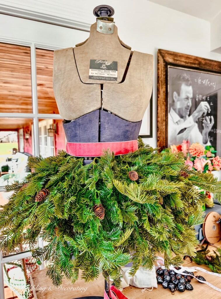 Vintage adjustable blue and gray bust form on tabletop styled with evergreens and a pink ribbon around the waist.