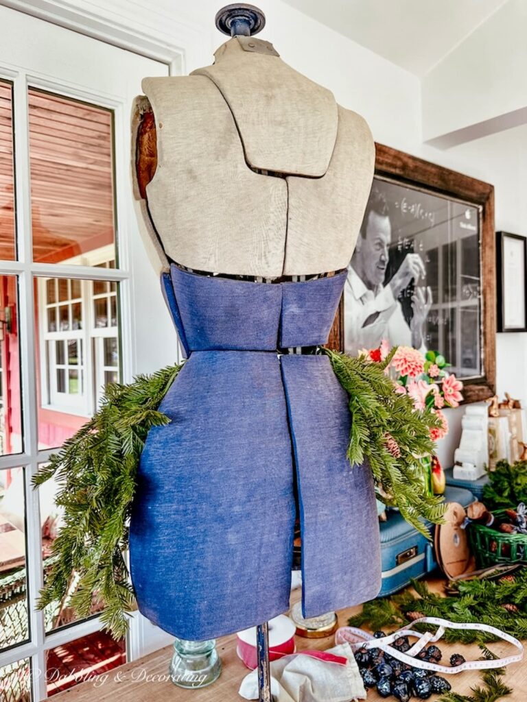 Vintage bust form in blue and gray on table with faux evergreens wrapped around half the waist.  Back side view.