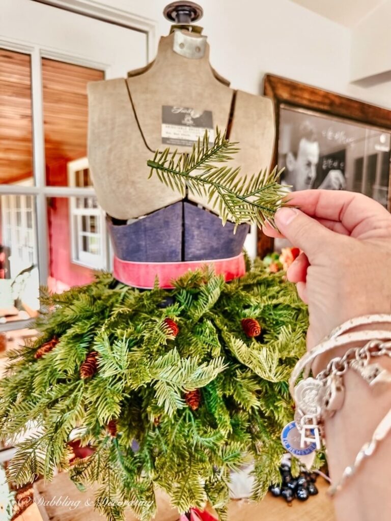 Vintage adjustable blue and gray bust form on tabletop styled with evergreens and a pink ribbon around the waist. and Hand holding smaller branch.