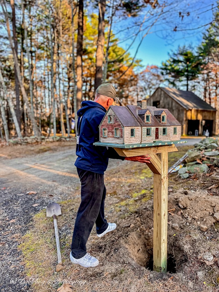 Man holding a unique mailbox post in the ground in front of house.