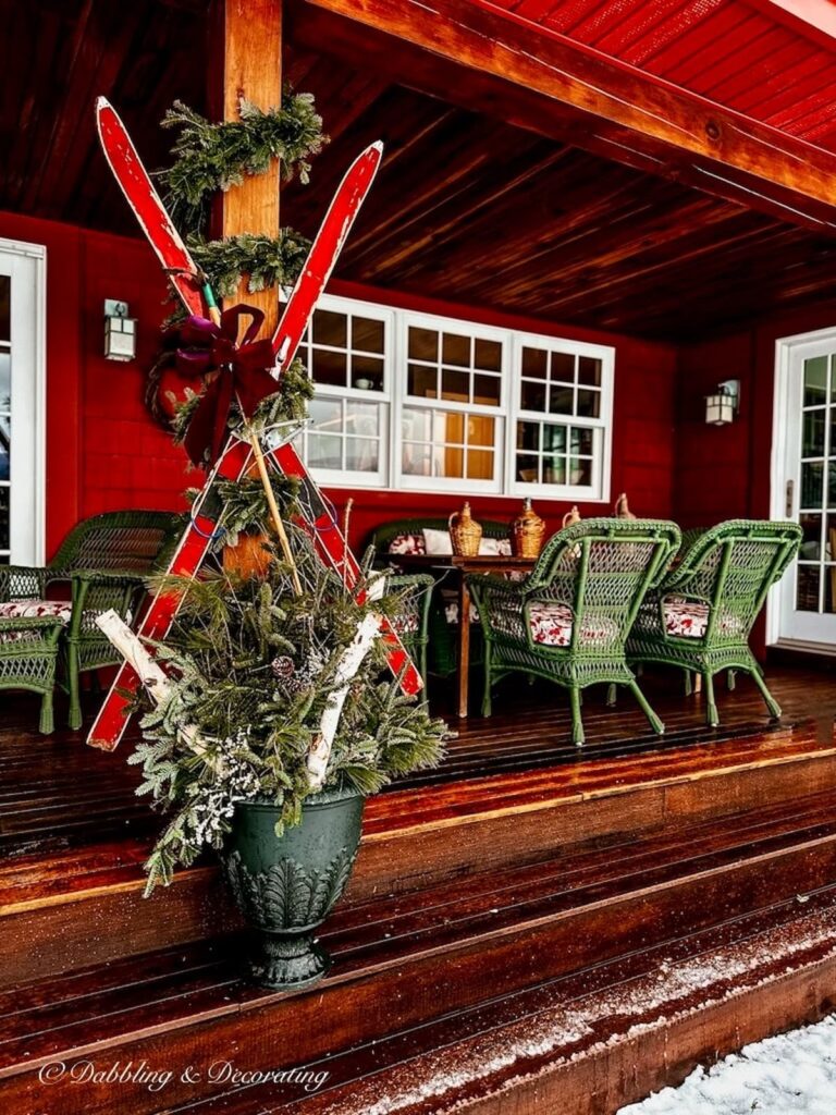 Vintage red criss crossed skis on Christmas wooden porch with evergreens and wicker seating.