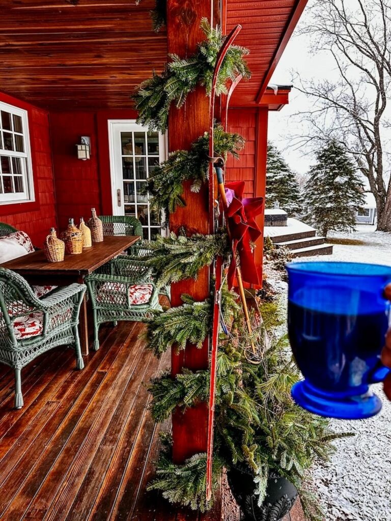 Vintage Christmas porch in red and green with vintage skis, demijohns and blue mug of coffee in the snow.