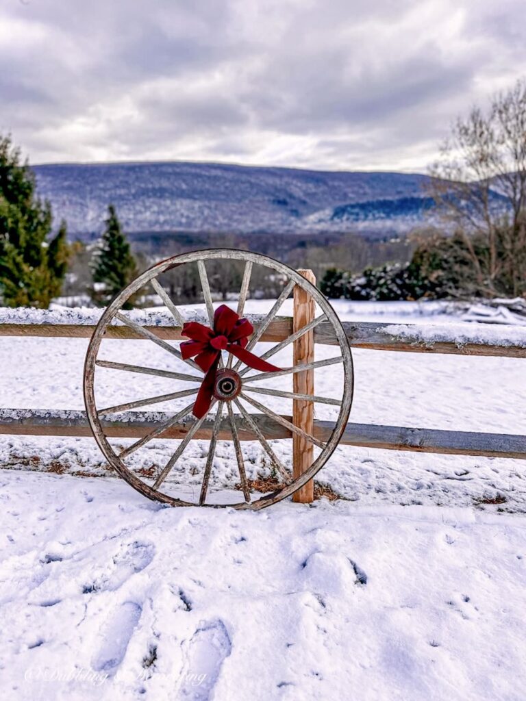 Vintage wooden wagon wheel with burgundy Christmas bow leaning on split rail fence in the mountains with snow.